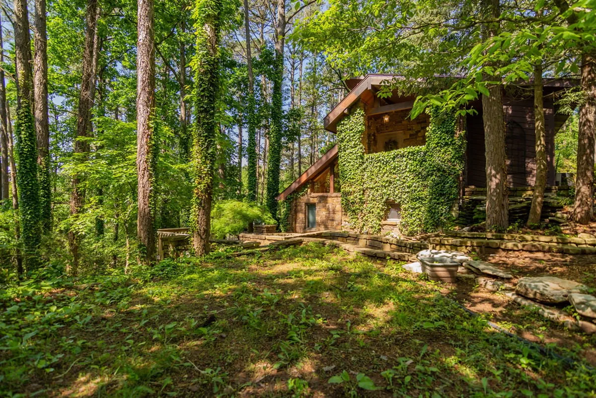 A tranquil exterior view of a wooden house nestled among lush greenery and tall trees, with stone steps leading to the entrance. The setting evokes a sense of peaceful seclusion and natural beauty, ideal for private getaways or family retreats.