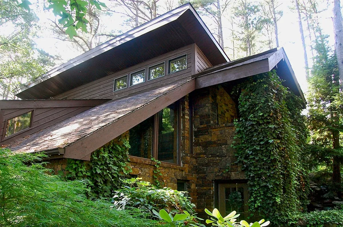 A scenic view of Tall Pines Sanctuary's exterior, featuring a sloping roof, expansive windows, and a stone facade nestled among lush greenery. The inviting entrance and well-maintained grounds convey a serene, natural environment perfect for exploring.