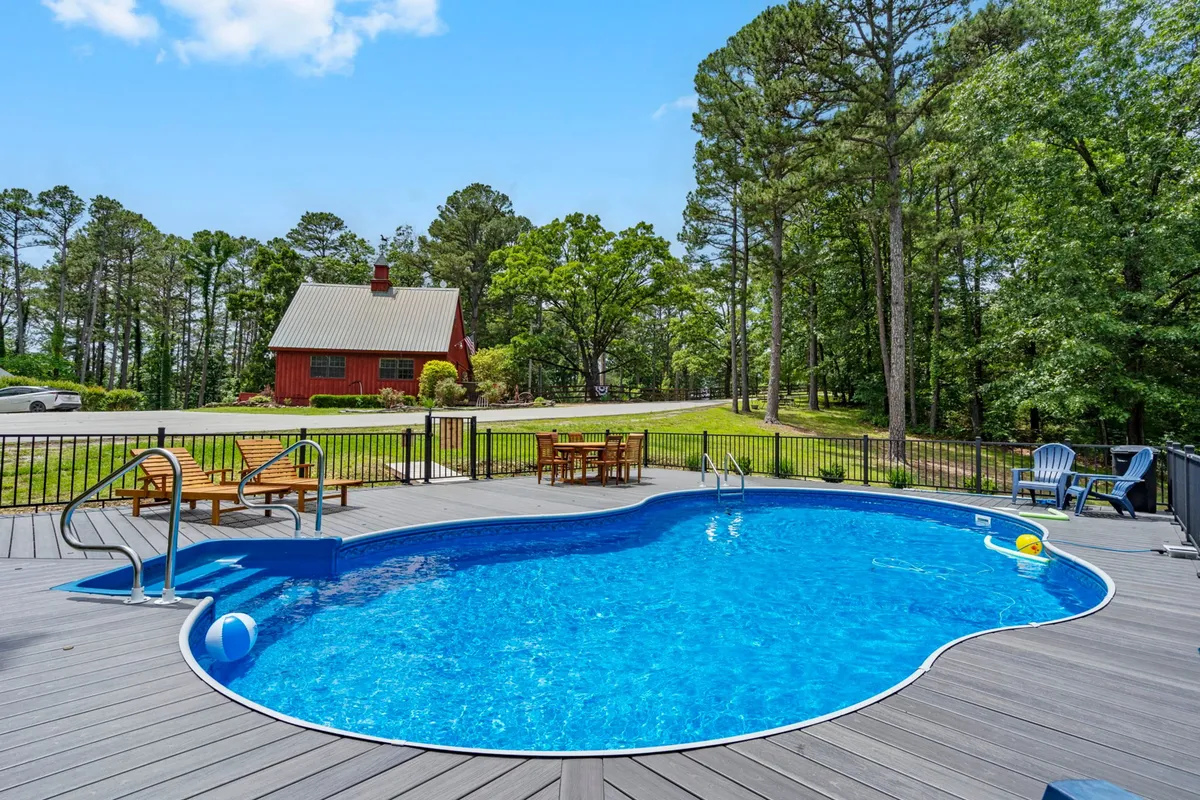 A sparkling swimming pool is surrounded by a red house, deck chairs, and lush trees, creating an inviting and leisurely outdoor gathering space. The pool is the centerpiece, perfect for sunny days and relaxed afternoons with family or friends.