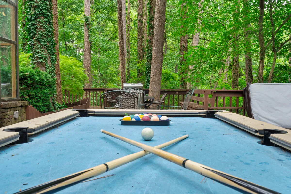 A spacious wooden deck surrounded by lush green foliage, featuring a pool table and multiple lounge chairs. The scene is lively, capturing the playful and social aspect of the deck, where guests can enjoy friendly competition or relax amidst nature.