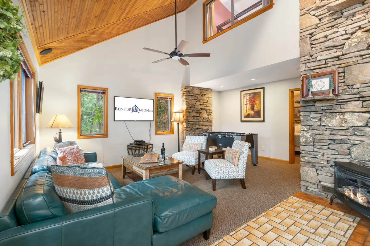 A family room with a teal couch arranged around a stone fireplace, beneath a wooden ceiling. Large windows frame the green forest outside, bathing the space in natural light and reinforcing the connection to nature.