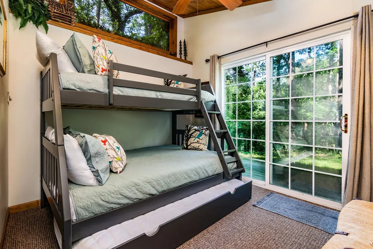 This image features a bright and inviting bunk room with bunk beds, large windows, and decorative pillows. The natural light streaming in creates a cozy, cheerful setting ideal for children or family stays.