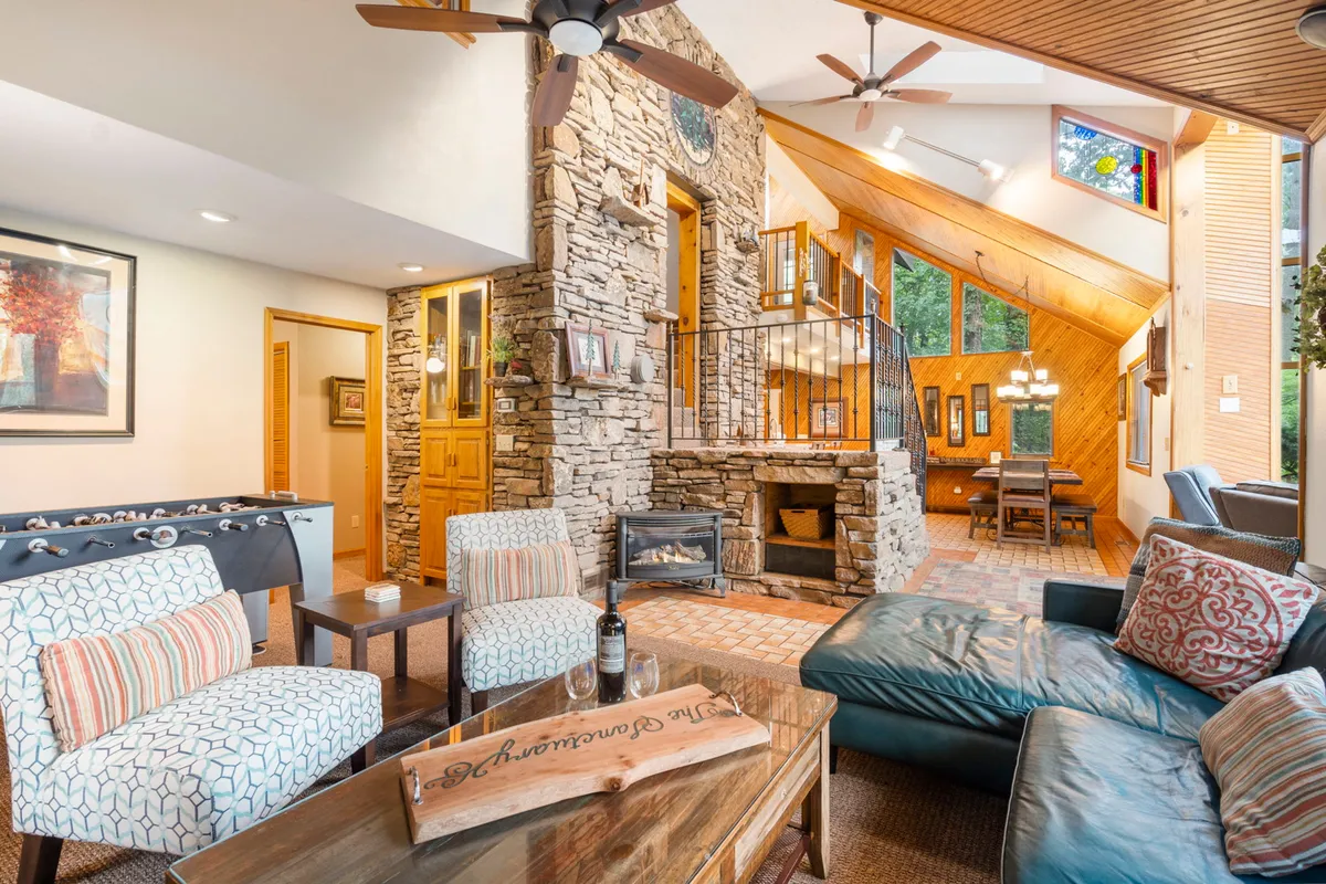 The image showcases a warm, inviting living area with a majestic stone fireplace, soaring wooden ceilings, expansive windows framing forest views, plush seating, and an adjoining dining space—creating a perfect setting for relaxation, game nights, and shared meals.