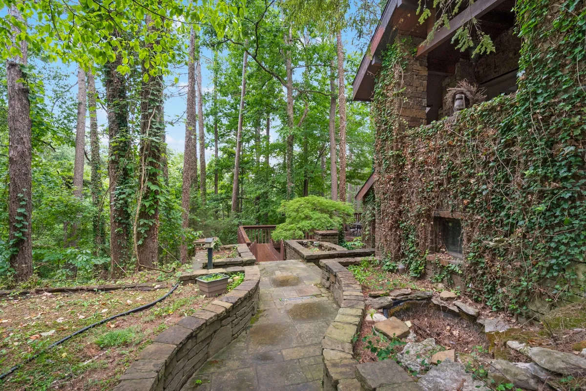 A serene exterior shot featuring an ivy-covered wall, stone pathway, wooden deck, and surrounding trees. The inviting space suggests places for morning coffee, evening storytelling, or simply soaking up the peaceful forest atmosphere.