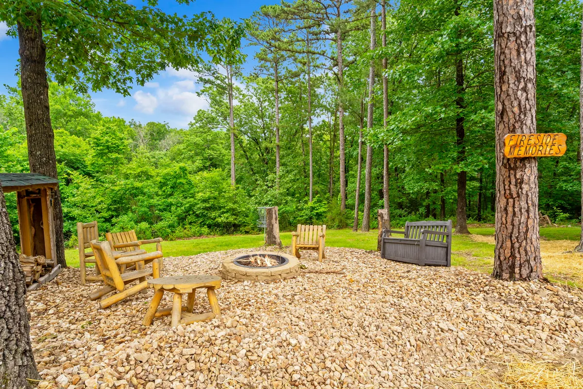 A cozy outdoor fire pit with wooden chairs is nestled among tall trees, with a welcoming sign and a tranquil forest backdrop. The setup invites guests to unwind, share stories, and enjoy evenings under the stars.