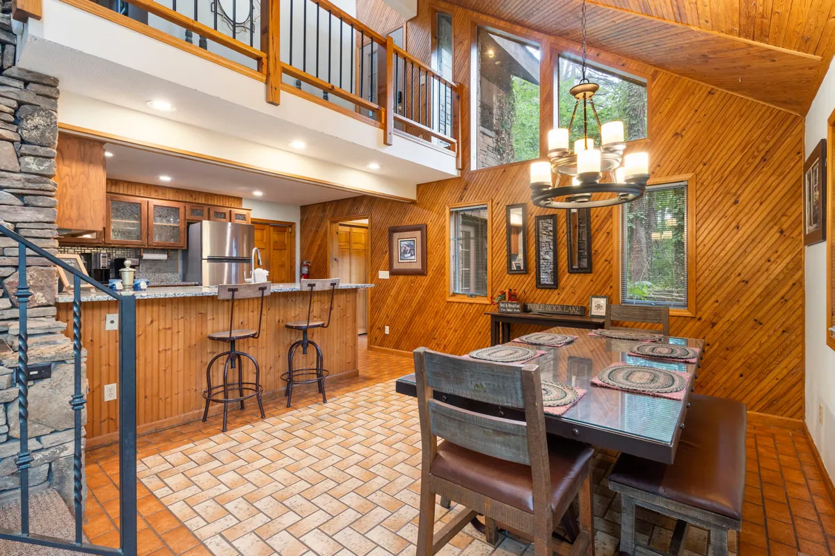 This image showcases an inviting open-concept space featuring a wooden dining table, a prominent stone fireplace, a kitchen island, and expansive windows that flood the room with natural light and woodland views. The layout connects the kitchen and dining areas, emphasizing togetherness and warmth.