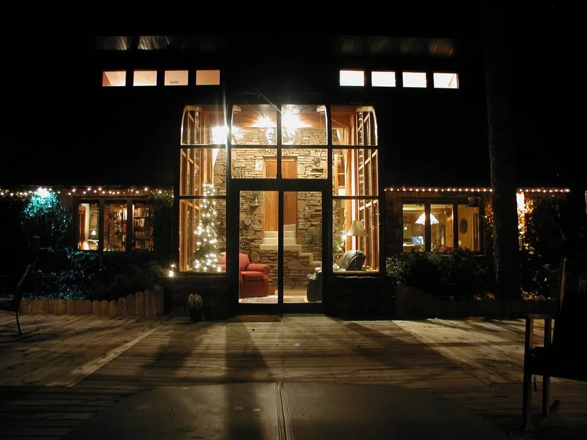 The image captures the large glass front of Tall Pines Sanctuary, with a striking stone entrance and decorative lights illuminating the exterior. Through the glass, a red chair is visible inside, hinting at the inviting interior. The setting is surrounded by natural elements, blending the architecture seamlessly with the wooded environment.