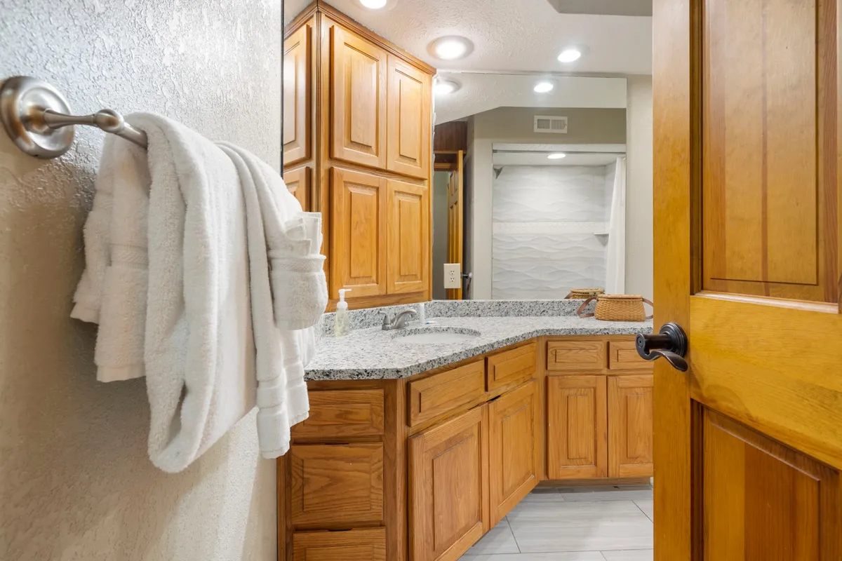 A calm and serene bathroom is depicted, with granite countertops, wooden cabinetry, neatly hung towels, and a large mirror reflecting the space. The ambiance is one of peaceful self-care and relaxation.