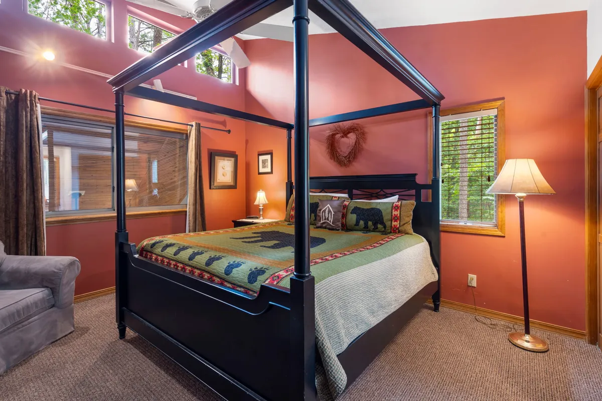This image presents a comfortable and serene bedroom featuring a four-poster bed, a decorative quilt, a sunlit window, a side lamp, and an armchair. The space feels warm, restful, and inviting.