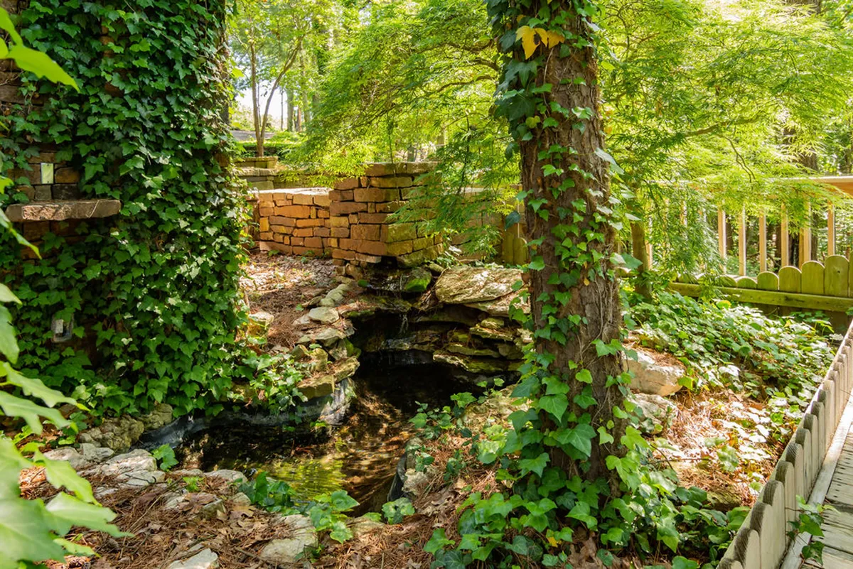 A serene exterior view of a stone structure accented by a lush green landscape and a tranquil water feature. The setting is surrounded by trees and vibrant foliage, embodying a peaceful connection to nature.